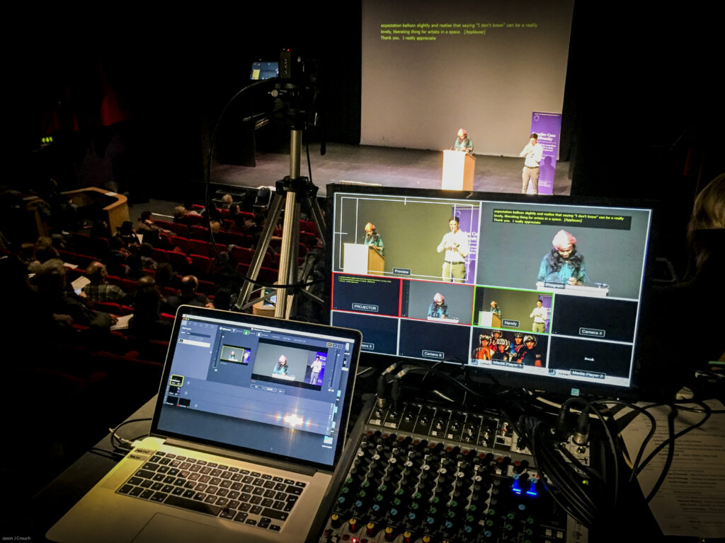 On a conference stage a speaker is stood behind a lectern. She is a tall dark skinned woman. To her left is a white male BSL interpreter stood in front of a roll-up banner. In the foreground is a collection of equipment being used to composite camera feeds of the speaker and the BSL interpreter. Captions are shown both projected above the speaker and on the live stream. 